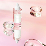 Un flacon compte-gouttes de sérum de soin, étiqueté « Centella », est posé sur une surface réfléchissante, entouré de sphères transparentes semblables à du gel. Le fond est un doux dégradé de teintes roses, créant une esthétique sereine et élégante. Dakar