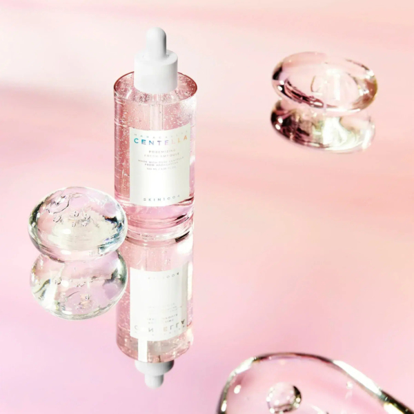 Un flacon compte-gouttes de sérum de soin, étiqueté « Centella », est posé sur une surface réfléchissante, entouré de sphères transparentes semblables à du gel. Le fond est un doux dégradé de teintes roses, créant une esthétique sereine et élégante. Dakar