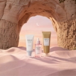 Trois produits de soin Skin1004 Coffret Illuminateur Anti Taches et Hydratant Édition Limitée 3 Pièces - deux tubes et un flacon compte-gouttes - sont posés sur du sable rose pâle avec, en arrière-plan, une arche de pierre, un ciel pastel et un croissant de lune. Dakar
