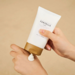 Une main presse un tube blanc de crème Centella sur le dos d'une autre main. La crème est distribuée par un bouchon beige, sur un fond beige uni. Dakar