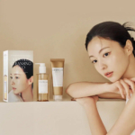 Une femme aux cheveux attachés est appuyée sur une table et regarde la caméra. À côté d'elle se trouvent des produits de soin, dont une boîte blanche avec une image, un flacon pompe et un tube, tous étiquetés « Centella ». Le fond est beige neutre. Dakar