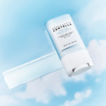 Un produit de soin bleu et blanc, étiqueté « Madagascar Centella HYALUCICA BLUE SERUM STICK », de SKIN1004, est présenté sur un fond de ciel doux et nuageux. Le stick est ouvert, révélant une petite partie du produit. Dakar