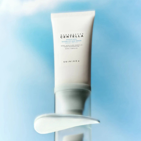 Un tube de sérum en feuille à la centella de Madagascar de SKIN1004 est positionné sur un fond bleu et blanc doux. Le tube est vertical et les informations sur le produit sont imprimées en noir. En dessous, une trace de sérum est visible sur une surface transparente. Dakar