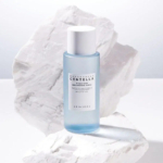 Un flacon bleu de tonique éclaircissant SKIN1004 Madagascar Centella Hyalu-Cica est posé sur un fond blanc texturé, évoquant des rochers. L'étiquette du flacon est visible, avec les détails du produit et la marque. Dakar
