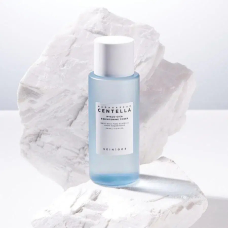 Un flacon bleu de tonique éclaircissant SKIN1004 Madagascar Centella Hyalu-Cica est posé sur un fond blanc texturé, évoquant des rochers. L'étiquette du flacon est visible, avec les détails du produit et la marque. Dakar