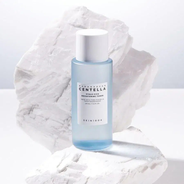 Un flacon bleu de tonique éclaircissant SKIN1004 Madagascar Centella Hyalu-Cica est posé sur un fond blanc texturé, évoquant des rochers. L'étiquette du flacon est visible, avec les détails du produit et la marque. Dakar