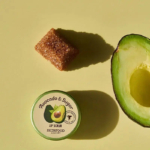 Un pot de gommage à lèvres à l'avocat et au sucre est posé sur un fond vert clair. À proximité, une moitié d'avocat coupée et un morceau de sucre roux projettent des ombres. Dakar