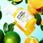Un flacon de nettoyant glycolique « Bye Bye Blemish » aux extraits d'agrumes est entouré de citrons verts frais et de tranches d'orange sur un fond bleu clair. Le flacon est jaune avec une étiquette blanche. Dakar