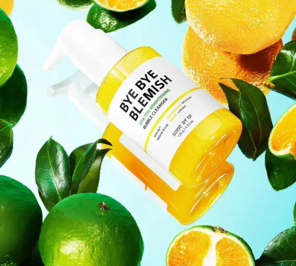 Un flacon de nettoyant glycolique « Bye Bye Blemish » aux extraits d'agrumes est entouré de citrons verts frais et de tranches d'orange sur un fond bleu clair. Le flacon est jaune avec une étiquette blanche. Dakar
