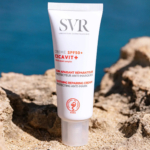 Un tube de crème réparatrice apaisante SVR Cicavit+ SPF 50+ est posé sur une surface rocheuse. Le tube est blanc avec un texte rouge et noir, et l'arrière-plan représente un paysage rocheux et sablonneux flou. Dakar