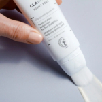 Une main tient un tube blanc de crème peeling de nuit et applique une fine couche de produit sur une surface lisse et grise. Le tube est orné d'un logo et d'un texte descriptif sur ses propriétés dépigmentantes, avec la mention « Fabriqué en France ». Dakar