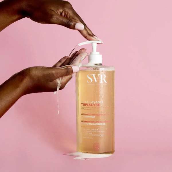 Mains pressant la pompe d'un grand flacon d'huile nettoyante SVR Topialyse, savonnant sur les doigts. Le flacon est transparent, avec un texte rose sur fond rose. Dakar