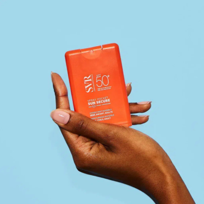 Une personne aux ongles rose clair tient un vaporisateur de crème solaire orange de poche avec un indice de protection solaire SPF 50+ sur fond bleu. Le produit est étiqueté « Sun Secure » et « SVR ». Dakar