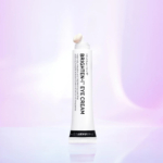 Un tube de crème contour des yeux Brighten-I de Beauty of Joseon se dresse sur un fond pastel flou. Le tube est blanc, avec un texte noir, et une petite quantité de crème est visible à l'ouverture. Dakar