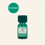 Un petit flacon vert de 10 ml d'huile d'arbre à thé The Body Shop, avec son bouchon vert, est présenté sur un fond crème clair. L'étiquette indique qu'il convient aux peaux à imperfections. Le texte « Iconique » dans un cercle vert apparaît en haut à gauche. Dakar