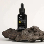 Un flacon de sérum Pore Minimizing 21, muni d'un bouchon compte-gouttes, est posé sur un morceau de roche volcanique poreuse. Le flacon noir est orné d'un texte jaune et blanc, soulignant les préoccupations concernant les pores et le sébum, ainsi que les bienfaits du resserrement des pores. Dakar