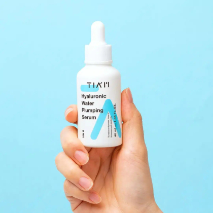 Une main tenant un flacon compte-gouttes blanc étiqueté « TIAM Hyaluronic Water Plumping Serum » sur fond bleu clair. Le flacon arbore un texte bleu et noir, avec des éléments minimalistes. Dakar