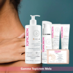 Gros plan sur une personne tenant une bouteille de lait ultra-hydratant Topicrem MELA. En arrière-plan, différents produits Topicrem sont exposés : lait hydratant, crème, pain de douceur, etc., tous étiquetés avec le logo de la marque Topicrem. Dakar