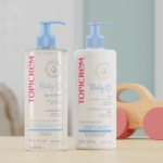 Deux flacons de produits Topicrem Bébé, un gel nettoyant et un lait ultra-hydratant, tous deux munis de pompes bleues, sont posés sur une surface en bois. Un jouet en bois à roues roses apparaît à l'arrière-plan à droite. Dakar