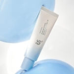 Un tube blanc et bleu clair de Beauty of Joseon Relief Sun Aqua Fresh Rice + B5 Fluide Solaire SPF50 au panthénol 50 ml avec un texte coréen est posé en diagonale sur un fond bleu et blanc doux, accentué par des formes rondes bleu clair. Dakar