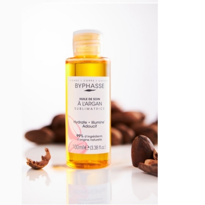 Un flacon d'huile de soin BYPHASE SUBLIMATRICE ARGAN 100 ml est présenté avec une étiquette transparente, entourée de noix d'argan concassées et d'une touche d'huile de sésame bio Waam. L'emballage transparent dévoile l'huile dorée à l'intérieur sur une surface blanche. Dakar