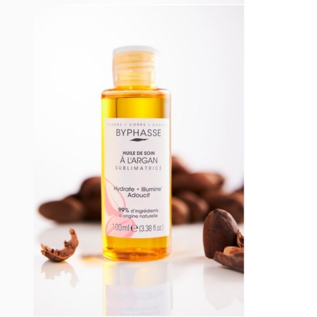 Un flacon d'huile de soin BYPHASE SUBLIMATRICE ARGAN 100 ml est présenté avec une étiquette transparente, entourée de noix d'argan concassées et d'une touche d'huile de sésame bio Waam. L'emballage transparent dévoile l'huile dorée à l'intérieur sur une surface blanche. Dakar