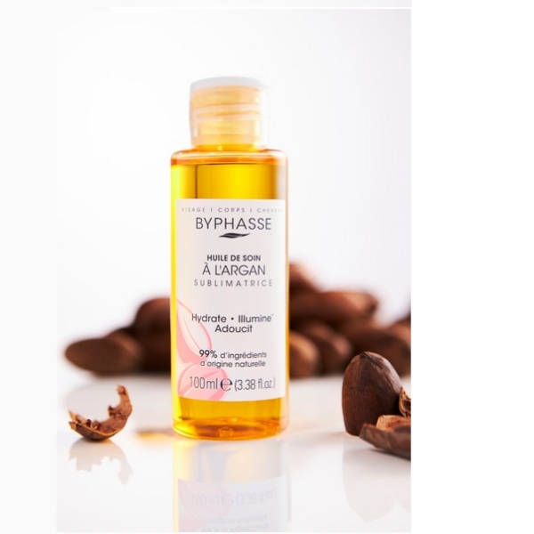 Un flacon d'huile de soin BYPHASE SUBLIMATRICE ARGAN 100 ml est présenté avec une étiquette transparente, entourée de noix d'argan concassées et d'une touche d'huile de sésame bio Waam. L'emballage transparent dévoile l'huile dorée à l'intérieur sur une surface blanche. Dakar
