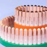 Un assortiment coloré de bâtons de fond de teint est disposé sur deux plateaux ronds, l'un orange, l'autre vert. Les bâtons présentent une palette de tons de peau, soigneusement disposés en motif ascendant sur un fond violet clair. Dakar