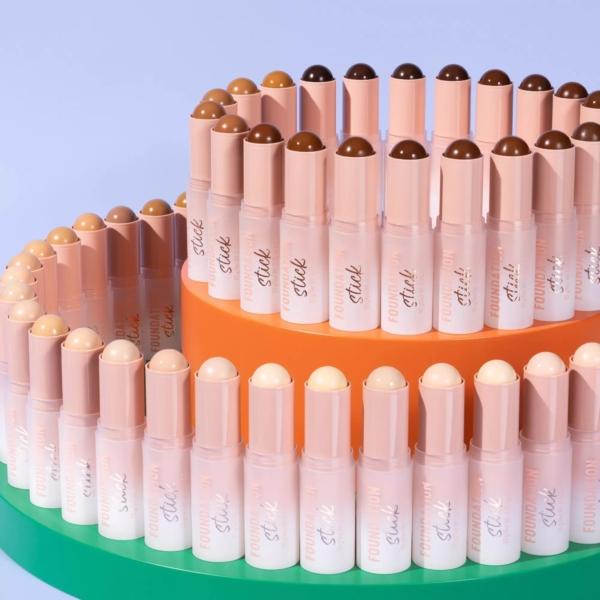 Un assortiment coloré de bâtons de fond de teint est disposé sur deux plateaux ronds, l'un orange, l'autre vert. Les bâtons présentent une palette de tons de peau, soigneusement disposés en motif ascendant sur un fond violet clair. Dakar