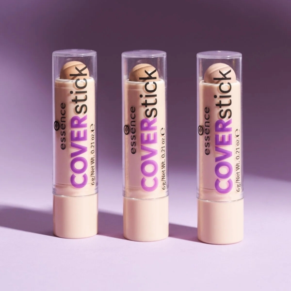 Trois bâtonnets Essence Cover Sticks se dressent sur un fond rose-violet. L'emballage est crème avec un texte violet, et les couvercles transparents révèlent le produit beige à l'intérieur. Les ombres s'étendent vers la gauche. Dakar