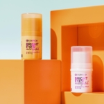 Deux sticks contour yeux anti-cernes Essence BRIGHT EYES, l'un orange et l'autre blanc, étiquetés « Effet éclaircissant et soin », reposent sur des blocs géométriques orange. Le fond vif, orné de formes et d'ombres, souligne l'élégance de ces essentiels pour le contour des yeux. Dakar