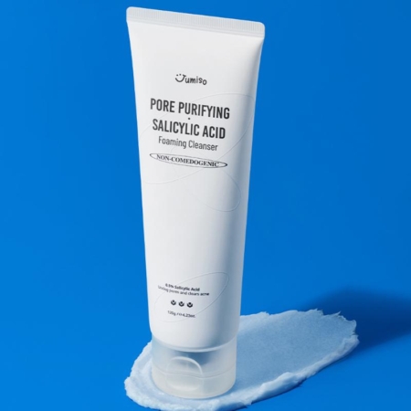 Un tube blanc de Jumiso Nettoyant Moussant Purifiant Anti Imperfections à l'Acide Salicylique 120 g se tient debout sur une tache de nettoyant avec un emballage et une marque minimes, sur fond bleu. Dakar