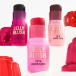 Quatre flacons de rouge à lèvres et de joues vibrants étiquetés « Jelly Blush » dans des tons de rouge, rose et violet sont disposés parmi des moules à gelée translucides de couleurs similaires, créant une composition ludique et colorée. Dakar