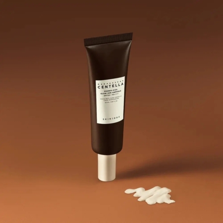 Un tube brun foncé de SKIN1004 Madagascar Centella Probio Cica SPF50 Ampoule Sérum Solaire 50 ml se tient debout sur une surface brune, avec une petite quantité de crème blanche pressée à côté. Dakar