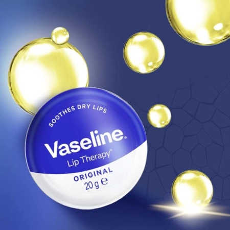 Une boîte de Vaseline Lip Therapy Original 17 g, étiquetée « Apaise les lèvres sèches », se dresse sur un fond bleu foncé avec des gouttelettes d'huile jaune translucide et un motif hexagonal subtil. Dakar