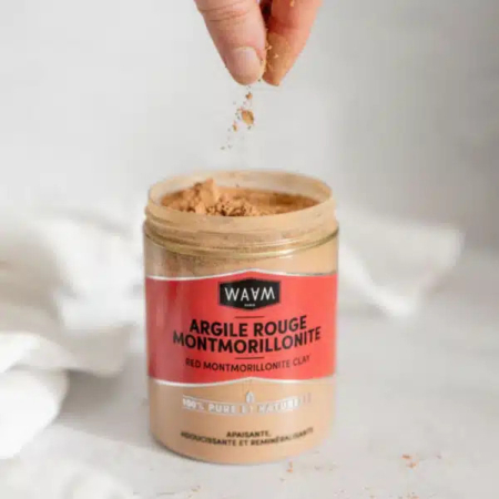 Une personne saupoudre de l'argile montmorillonite brun rougeâtre provenant d'un pot étiqueté « WAVM Argile Rouge Montmorillonite ». L'argile est commercialisée comme 100 % pure et naturelle. Le fond est blanc et légèrement flou. Dakar