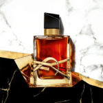 Un flacon de parfum en verre contenant un liquide ambré est posé sur une surface en marbre blanc. Il est doté d'un bouchon rectangulaire noir et d'une collerette dorée ornée d'un élément décoratif doré. L'arrière-plan de l'image ajoute une touche d'élégance. Dakar
