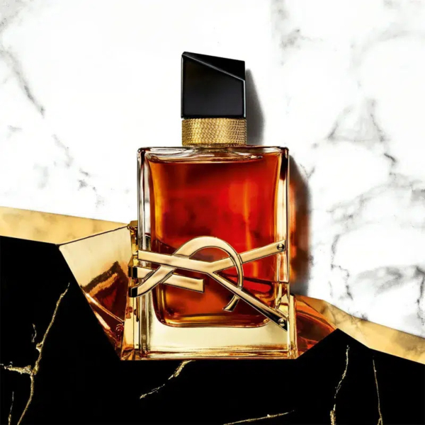Un flacon de parfum en verre contenant un liquide ambré est posé sur une surface en marbre blanc. Il est doté d'un bouchon rectangulaire noir et d'une collerette dorée ornée d'un élément décoratif doré. L'arrière-plan de l'image ajoute une touche d'élégance. Dakar