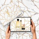 Une personne tient un coffret cadeau YSL sur un fond marbré veiné d'or. Le coffret contient un flacon de parfum, un tube fin, probablement destiné à un parfum ou à un soin, et un rouge à lèvres. Le couvercle et le fond du coffret présentent des motifs marbrés aux lignes dorées. Dakar
