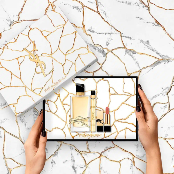 Une personne tient un coffret cadeau YSL sur un fond marbré veiné d'or. Le coffret contient un flacon de parfum, un tube fin, probablement destiné à un parfum ou à un soin, et un rouge à lèvres. Le couvercle et le fond du coffret présentent des motifs marbrés aux lignes dorées. Dakar