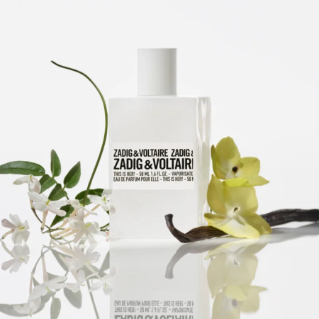Un flacon du parfum « Zadig & Voltaire This Is Her ! » orné d'un texte noir audacieux sur un flacon rectangulaire transparent. Autour de lui, de délicates fleurs de jasmin blanc, une fleur jaune et deux gousses de vanille, dont la surface se reflète délicatement. Dakar