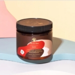 Un pot d'Asiam Cache-cache à la noix de coco 16 onces avec un couvercle noir et une étiquette rouge, beige et marron repose sur une surface pastel avec des formes abstraites en arrière-plan, soulignant son soin nourrissant. Dakar
