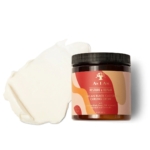Un pot de Crème à Friser à l'Huile de Ricin Noir de Jamaïque Asiam 227 g se trouve à côté d'une tache de crème blanche sur un fond blanc, tous deux présentés avec un emballage élégant et un étiquetage élégant. Dakar