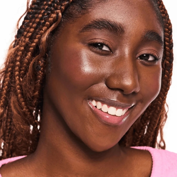 Une femme aux cheveux tressés arbore un sourire éclatant, révélant une peau radieuse et éclatante, sublimée par le stick blush et bronzeur Baby Got 40 Sweets & Roses d'Essence Cosmetics (5,5 g). Elle porte un haut rose clair et se tient debout sur un fond blanc. Dakar