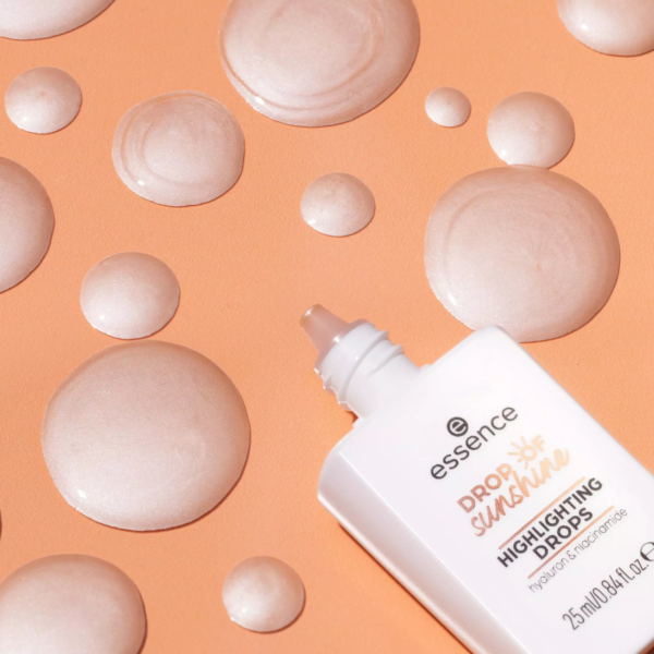 Un flacon d'Essence Gouttes Illuminatrices Universelles à base d'Acide Hyaluronique et de Niacinamide 25 ml est posé sur un fond pêche, entouré de gouttes artistiques de son liquide chatoyant. Dakar