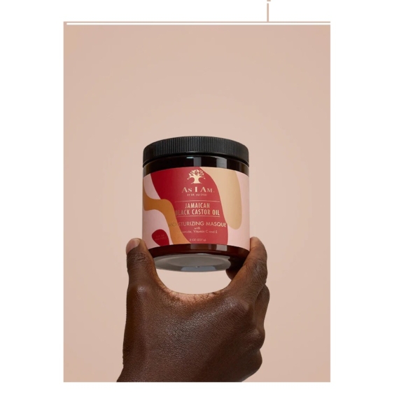 Une main tient un pot d'Asiam Masque capillaire à l'huile de ricin noire jamaïcaine 227 g sur un fond uni beige, évoquant le soin nourrissant du Masque Capillaire à la Biotine Soin Réparateur ADVANCED CLINICALS. Dakar