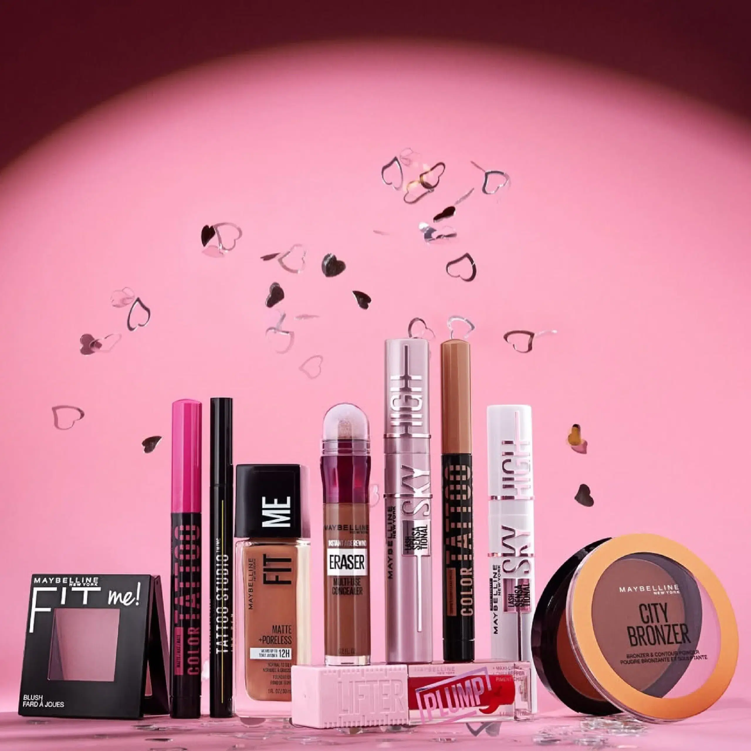 Une collection de produits de maquillage Maybelline d'Univers Cosmetix, comprenant fond de teint, anti-cernes, mascara, brillant à lèvres et poudre bronzante, disposés sur une surface rose avec des confettis en forme de cœur tombant en arrière-plan. Dakar
