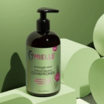 Un flacon pompe de 355 ml d'après-shampoing MIELLE Organics au Romarin est posé sur une plateforme géométrique vert clair sur fond vert. À associer au shampoing fortifiant MIELLE Organics pour des cheveux plus forts et plus sains, enrichi en biotine. Dakar