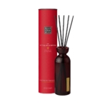 Un récipient cylindrique rouge étiqueté « Rituals Mini Bâtonnets Parfumés 70 ml » se trouve à côté d'un flacon en verre côtelé foncé avec des bâtonnets de roseau noirs, servant de diffuseur de parfum. Dakar