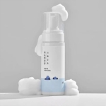 Un flacon blanc de mousse nettoyante Dokdo Round Lab 1025 de 150 ml, muni d'une pompe, libère une mousse blanche et onctueuse. Des motifs de montagnes bleues et un texte coréen ornent le flacon, entouré de mousse sur une surface gris clair. Dakar
